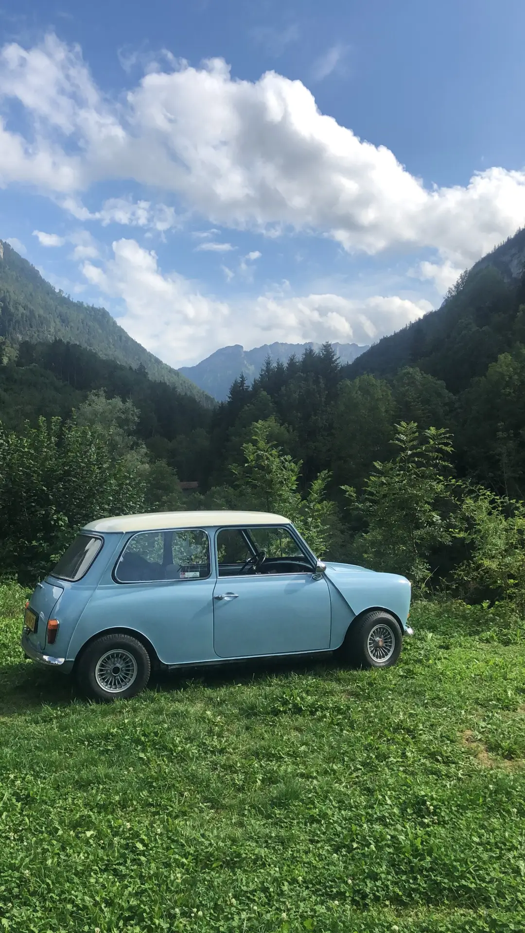 A light blue Austin Mini on a grass field in between mountains