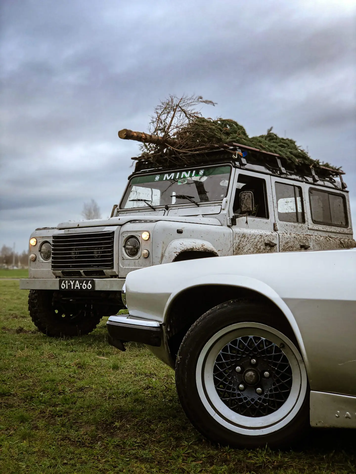 The wheel of a Jaguar XJS V12 with in the back the Brooks company car, the white Land Rover with a Christmas tree on top.